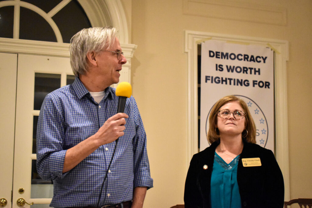 A man with white hair and a blue shirt speaks into a yellow microphone, standing next to a woman with brown hair and glasses at a Democrats of Davidson meeting. 