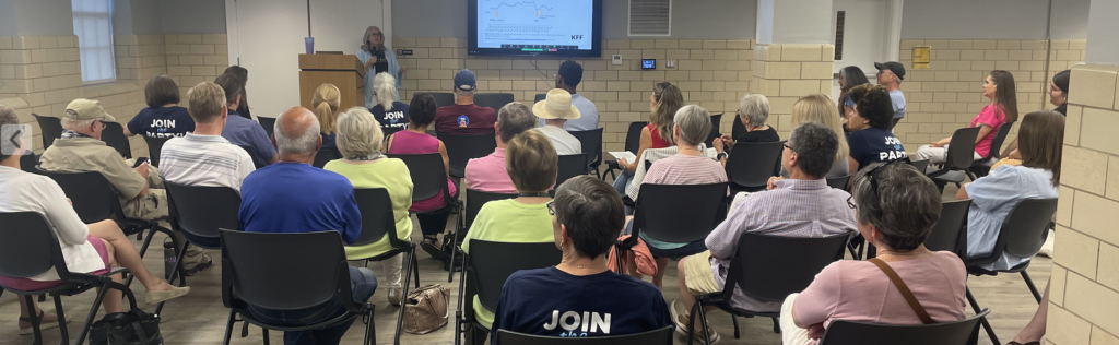 A group of 28 people sit down facing a woman speaking at a podium. The speaker is next to a television displaying information that relates to her speech. 