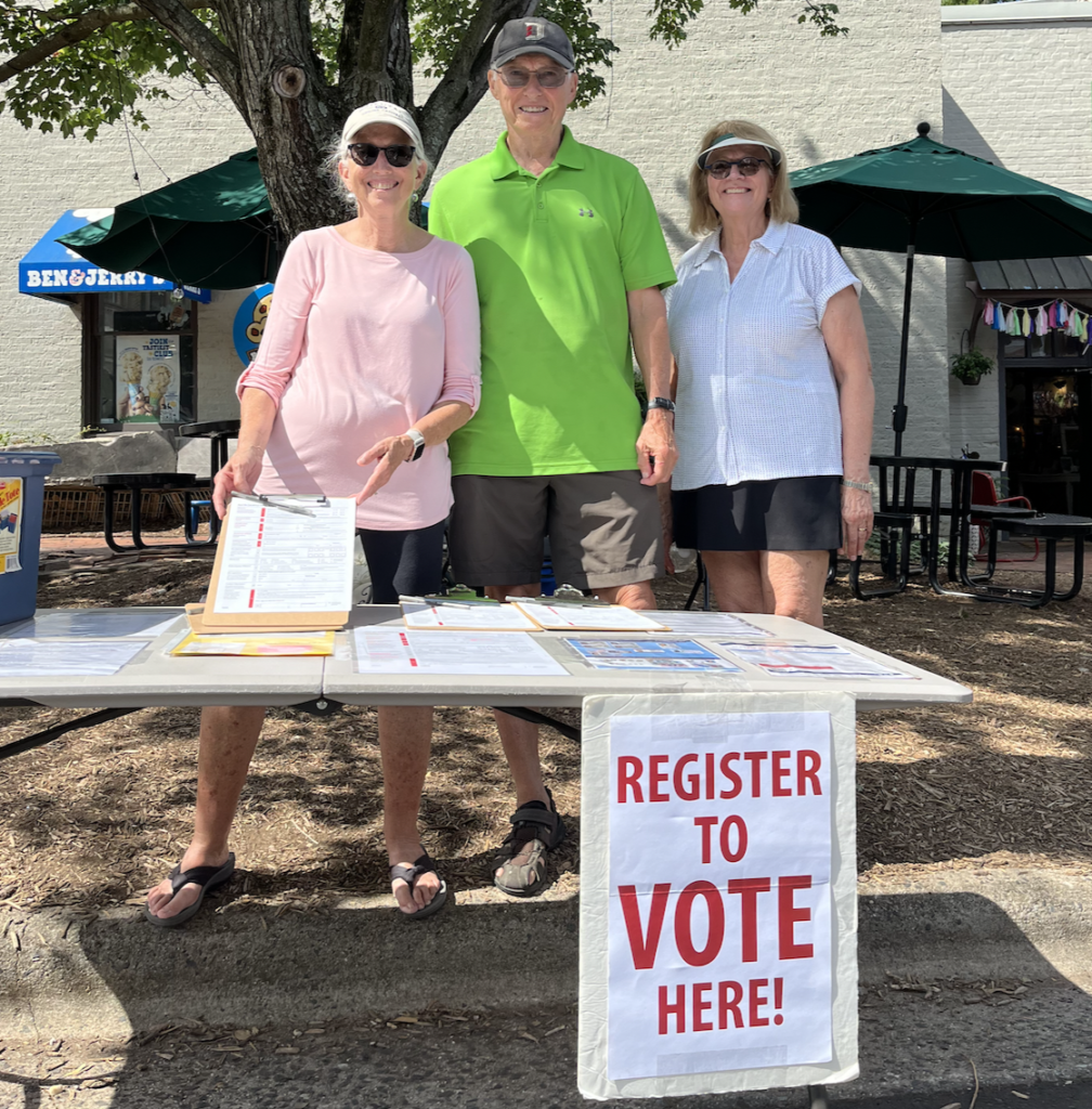 Two women and one man stand outside at a table with voter registration forms. They stand behind a white sign that says "Register to Vote here!" in red letters. 